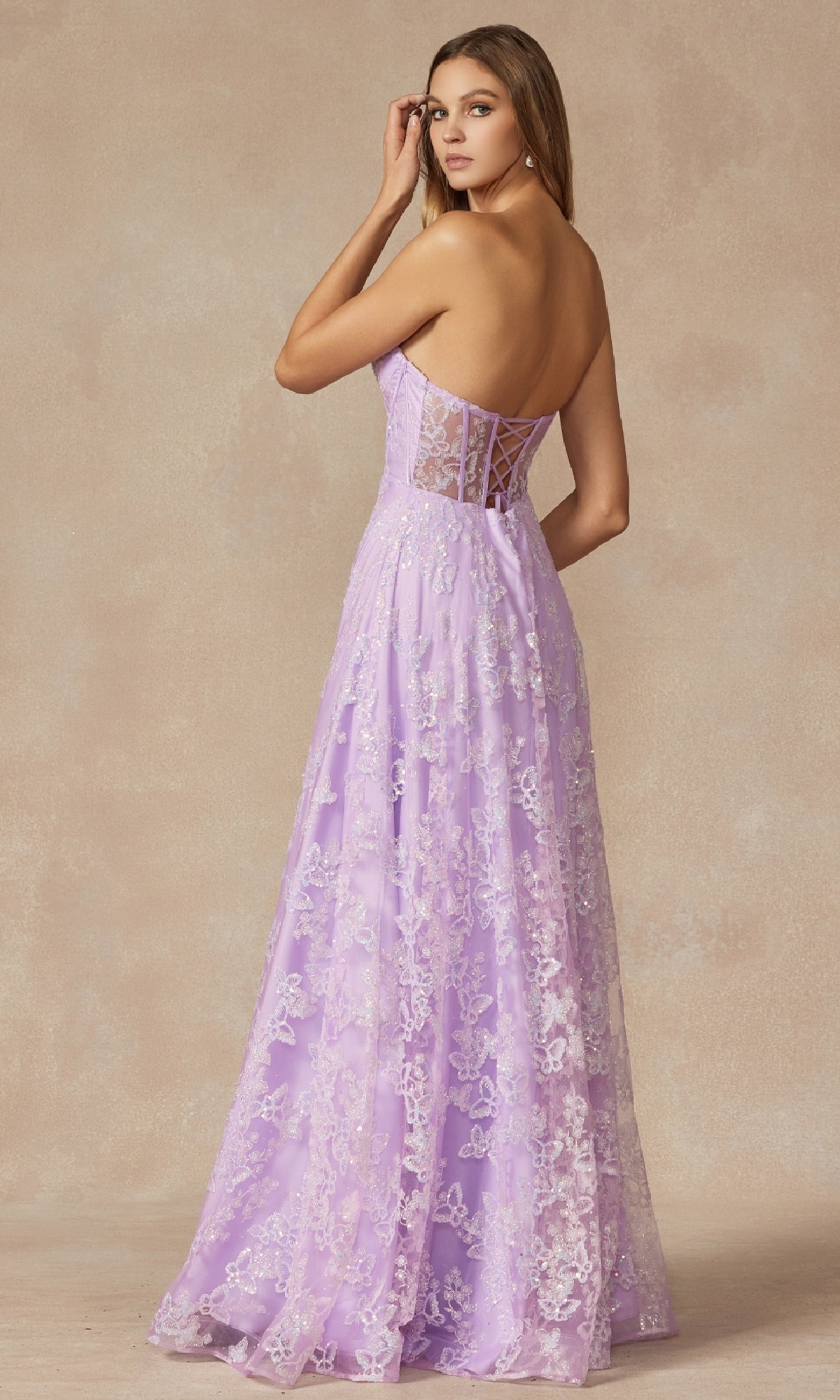 A woman models the Juliet 2403 long prom dress featuring a strapless lavender lace corset bodice and sheer lace back, as she looks over her shoulder against a neutral background.